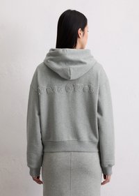 Sweat-shirt gris avec capuche, présentant une texture douce, une coupe ample et un logo brodé dans le dos. Jupe grise assortie visible en dessous.