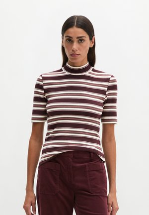 Vrouw met stijl donker haar draagt een bordeauxrood-wit gestreept kortemouws coltruitje en een bordeauxrode broek met steekzakken.