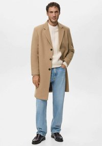 Abrigo de lana beige con botones negros, suéter crema de punto trenzado, vaqueros azules y zapatos de cuero negros, posando frente a un fondo neutro.