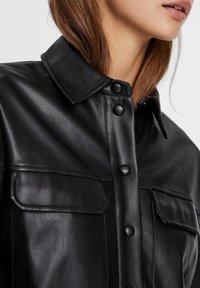 Chemise en cuir noir avec fermeture à boutons, grand col et deux poches poitrine. Présente une texture lisse et des boutons de tailles variées.