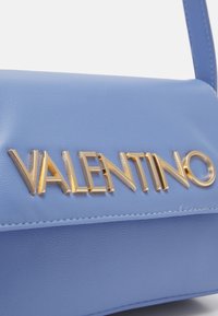 Ljusblå läderhandväska med en framträdande guld "VALENTINO"-logotyp. Slät textur med en strukturerad design och en stilren axelrem.