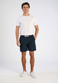 T-shirt bianca abbinata a pantaloni corti a righe blu scuro. I pantaloni hanno una vita elasticizzata e un cordoncino, e il modello indossa scarpe da ginnastica bianche.
