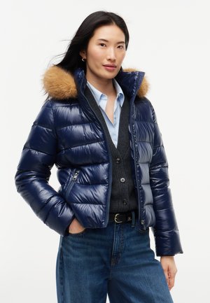 Mujer que lleva una chaqueta acolchada azul marino con capucha con ribete de piel sobre un cárdigan oscuro y camisa azul clara, con vaqueros azules, de pie frente a un fondo blanco.