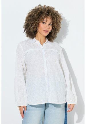 Donna con capelli ricci che indossa una camicia bianca a maniche lunghe con bottoni e un delicato motivo floreale, abbinata a jeans azzurri, su uno sfondo uniforme.