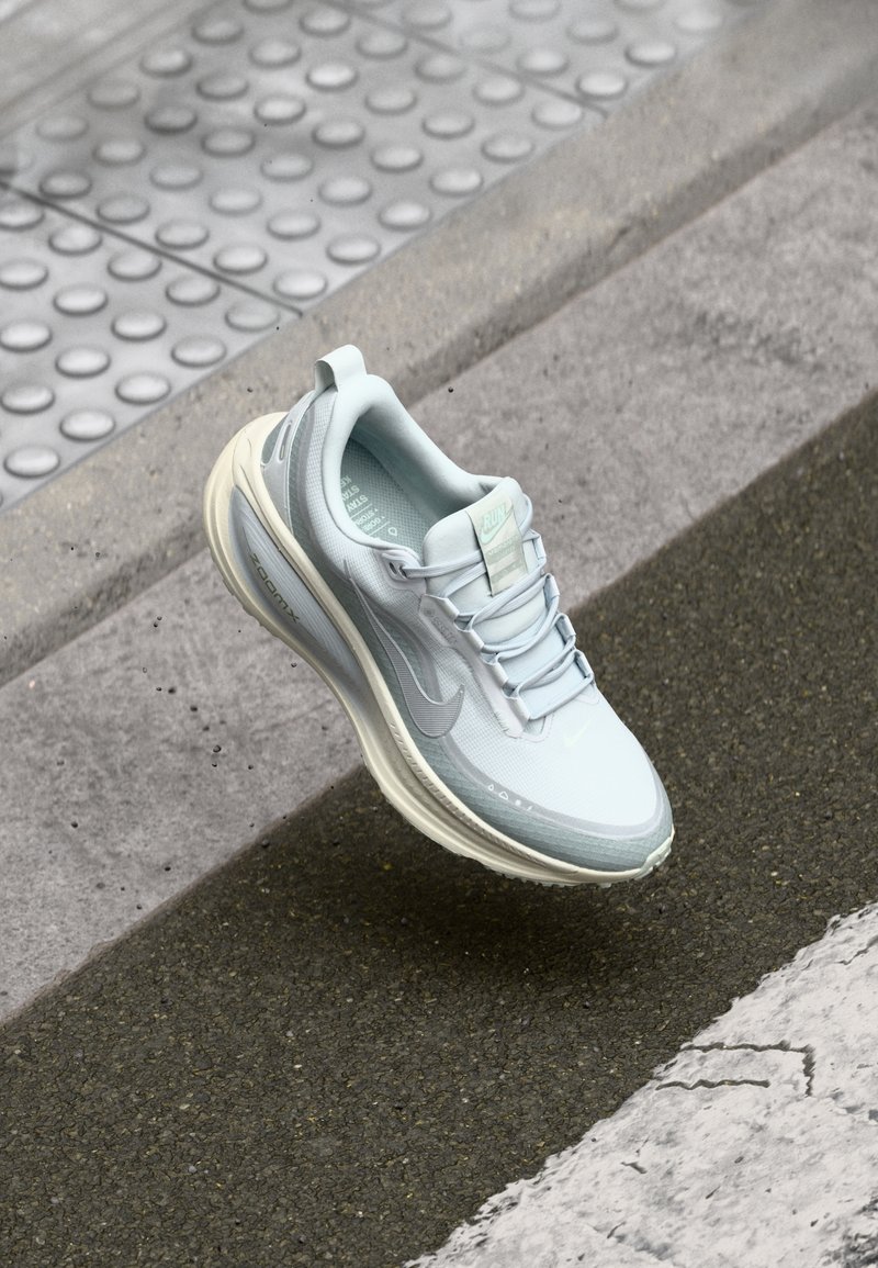 Chaussure de sport gris clair avec une semelle blanche, suspendue au-dessus d'un trottoir texturé et d'un bord de trottoir avec des pavés tactiles.