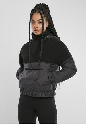Svart genserjakke med en fuzzy fleece-overdel og glatt nylon-nederdel. Har høyt krage, elastiske mansjetter og snor i sømmen.