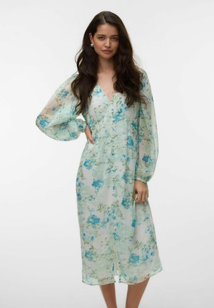 Nő könnyű, áttetsző midi ruhában, kék és zöld virágmintával, V-nyakkivágással, hosszú fodros ujjal és elöl gombos részletekkel.