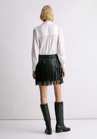 Zwarte leren rok met franjes, gecombineerd met een witte blouse met lange mouwen. Model draagt knielange zwarte laarzen met een ritsdetail.