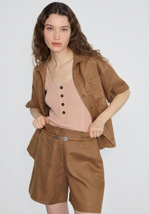 Giovane donna con capelli lunghi e ricci che indossa pantaloncini marroni, una camicia marrone a maniche corte abbinata con bottoni e una canottiera rosa pallido.