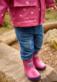 Botas de lluvia rosas con suelas azul marino, etiquetadas "JoJo Mama Bébé Splash," combinadas con unos vaqueros azules arremangados y una chaqueta de flores a juego.