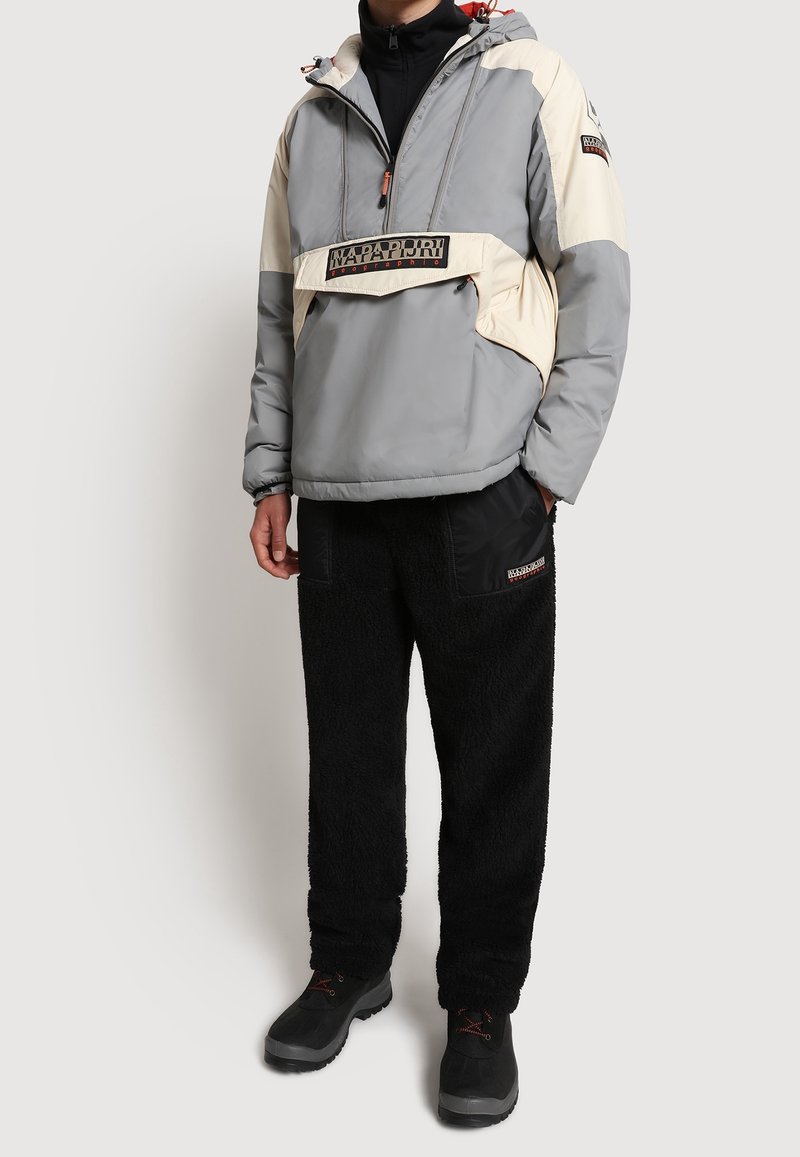 Personne portant une veste à capuche grise et beige avec poche avant, un pantalon en polaire noir et des bottes noires sur un fond uni.