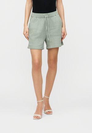 Kobieta ubrana w jasnoniebieskie, teksturowane szorty z troczkiem i beżowe sandały na wysokim obcasie na jednolitym tle.