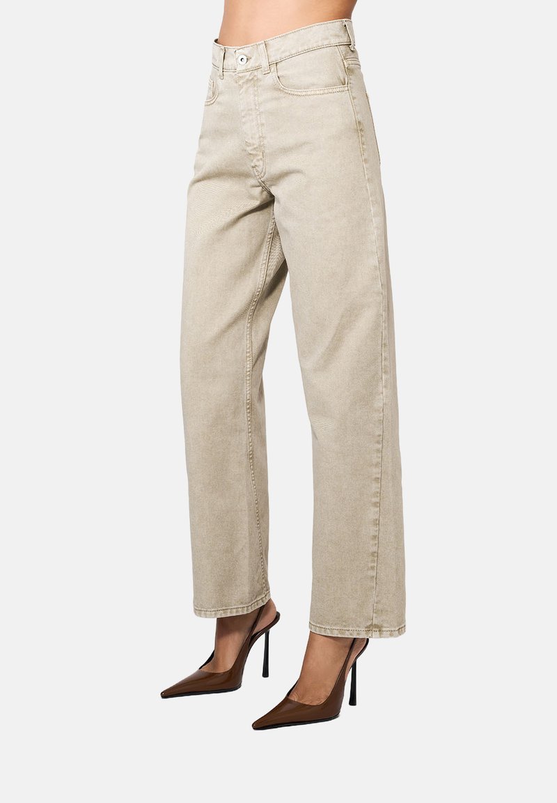 Beežid kõrge vöökoht, sirged jalgevahe teksad, mille jalas on tumepruunid terava ninaga slingback kontsad heledal taustal.