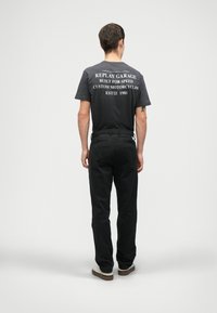 Άνδρας γυρισμένος πλάτη, φορώντας μαύρο T-shirt με λευκό κείμενο σχετικά με προσαρμοσμένες μοτοσυκλέτες, μαύρο παντελόνι και λευκά αθλητικά παπούτσια, σε απλό φόντο στο στούντιο.