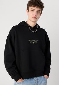 Schwarzer Hoodie aus Baumwollmischung mit einer vorderen Tasche und einem dunkelgrün gestickten Logo mit Flügel-Details. Sichtbare Kettenhalskette.