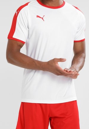 Man draagt een wit Puma sportshirt met rode accenten en rode shorts, houdt de rechterhand met de palm naar boven boven de linkerhand.