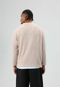 Beige, pluizige trui met een ontspannen pasvorm, lange mouwen, ronde hals en een gestructureerd oppervlak; gedragen over een wit shirt en zwarte broek.