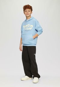Lichtblauw hoodie sweatshirt met grafische print, voorzak; gecombineerd met zwarte wijde broek en witte sneakers. Effen achtergrond.