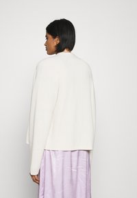 Witte gebreide cardigan met een relaxte pasvorm, ronde halslijn en afgezakte schouders, gedragen over een glanzende lavendel maxi-rok.