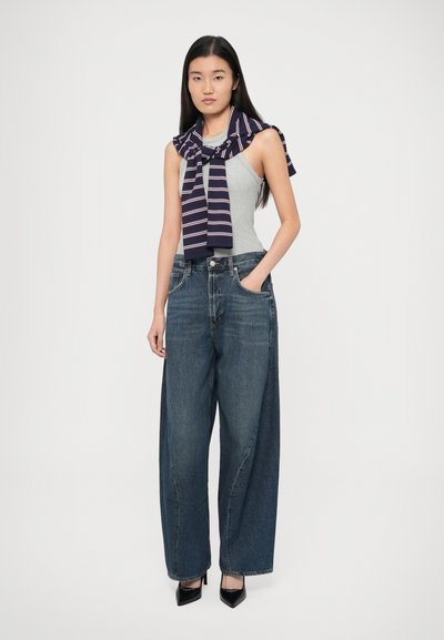 Γκρι ανοιχτό αμάνικο τοπ συνδυασμένο με φαρδιά, σκούρα μπλε τζιν. Μια μπλε ναυτική ριγέ εσάρπα δεμένη γύρω από τους ώμους. Μαύρα παπούτσια με ψηλό τακούνι.