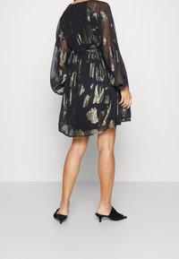 Svart, genomskinlig klänning med guldiga, abstrakta mönster, långa ärmar, markerad midja och flytande kjol. Matchad med svarta, öppna klackskor utan bakrem.