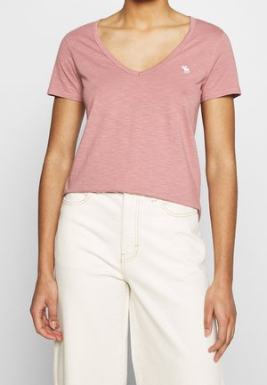 Vrouw draagt een stoffig roze V-hals T-shirt met een klein wit elandlogo en een hoog taille off-white broek, handen ontspannen langs haar zij.