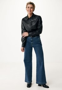 Camicia in pelle nera con colletto chiodato e tasca, abbinata a jeans in denim blu a gamba larga e stivali cuissard neri con tacco. Sfondo semplice.