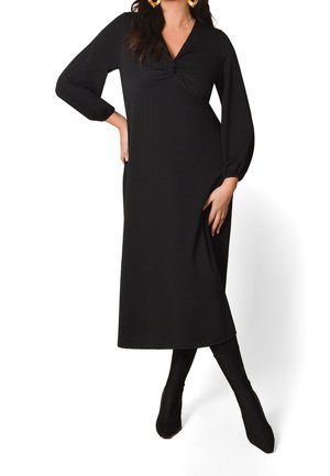 Vrouw draagt een zwarte midi-jurk met lange mouwen en een geplooide halslijn, gecombineerd met zwarte aansluitende laarzen en grote gouden creolen.