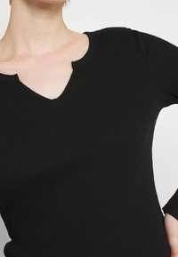 Schwarzes, geripptes Langarmshirt mit V-Ausschnitt. Der Stoff ist weich, und das Design bietet eine figurbetonte Silhouette mit sichtbaren Nähdetails.