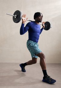 Ein Athlet führt einen Squat mit einer Langhantel aus. Er trägt ein langärmliges, navyfarbenes Oberteil, türkisfarbene Shorts mit Tierdruck und schwarze Sportschuhe mit goldenen Akzenten.