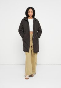 Sort quiltet lang frakke med hætte, der har trykknaplukninger og to forlommer, parret med beige cargo-bukser og beige sneakers.
