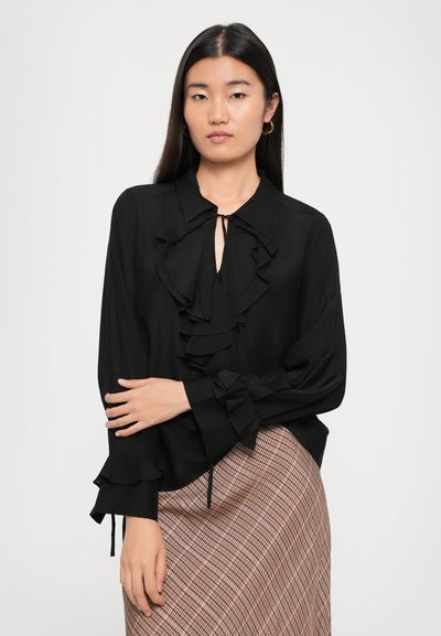 Black blouse with a ruffled front, soft fabric, and long sleeves featuring ruffled cuffs. Worn with a patterned skirt.