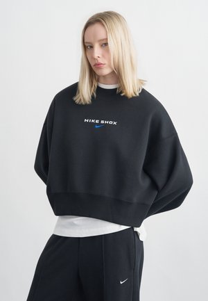Femme blonde portant un sweat-shirt Nike Shox noir surdimensionné et un pantalon Nike noir, debout devant un fond clair uni.