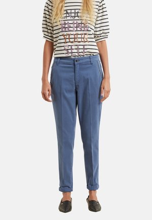 Frau trägt blaue, umgeschlagene Hosen, schwarze spitze Loafers und ein weiß gestreiftes Hemd mit dem Schriftzug "NICE BEING YOUR SELF" in Pailletten.