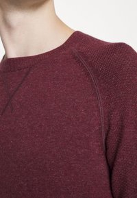 Burgunderfarbener Strickpullover mit Rundhalsausschnitt, der strukturierte Schultern und einen glatten Körper aufweist, aus weichem Material gefertigt.