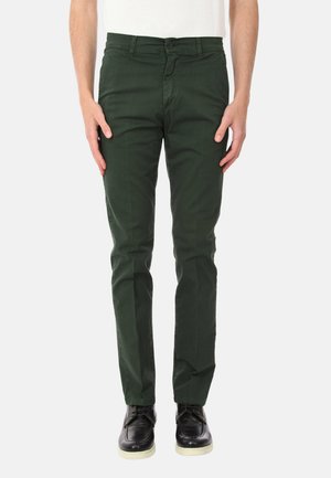 Uomo che indossa pantaloni slim fit verde scuro e scarpe di pelle nere, in piedi davanti a uno sfondo bianco semplice.