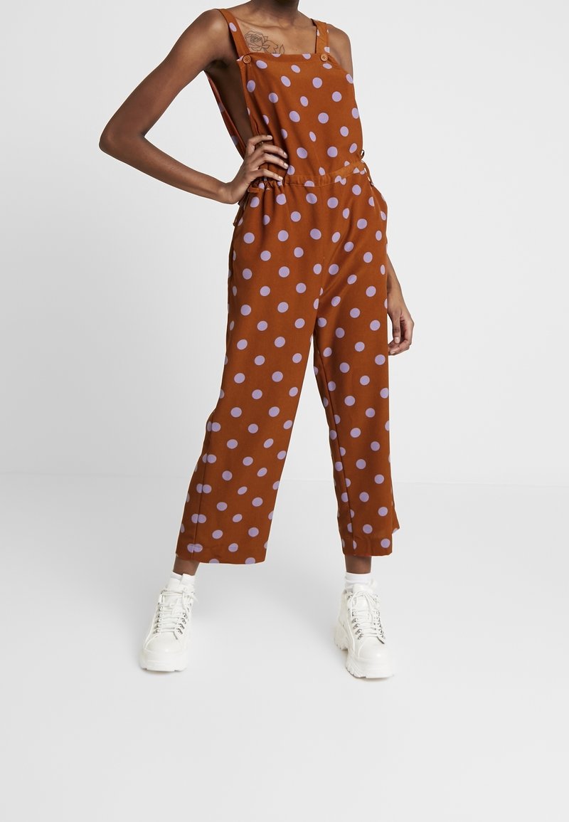 Brązowy kombinezon w kropki z luźnym krojem, szerokimi nogawkami, regulowanymi ramiączkami i ściągniętą talią, zestawiony z białymi trampkami.