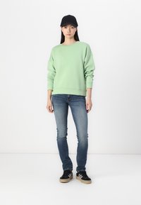 Sweat-shirt vert clair à manches amples, associé à un jean skinny bleu et des baskets noires. Une casquette noire complète cette tenue décontractée.