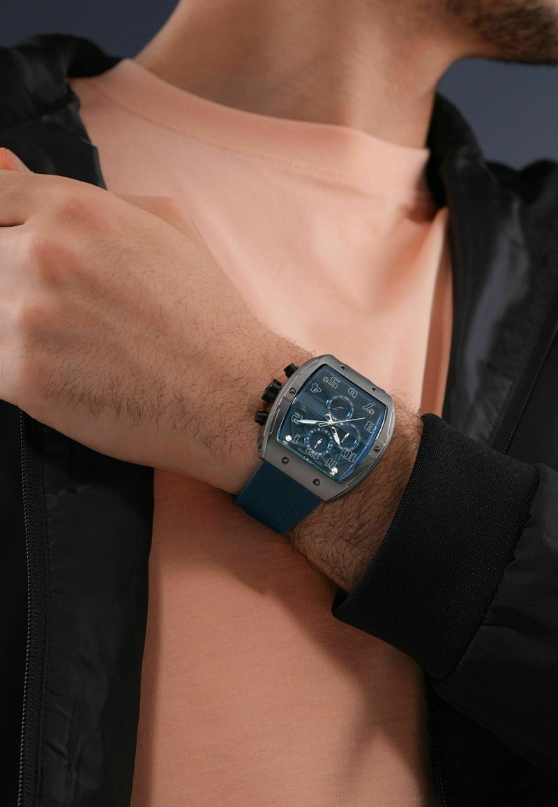 Blauwe siliconen horloge met een grijze metalen behuizing, hoekige vorm, transparante wijzerplaat en meerdere wijzerplaten en chronograaffuncties.