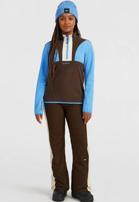 Pull-over bleu clair et marron avec col zippé et accents beiges, associé à un pantalon marron avec une bande beige sur le côté. Bottes noires et bonnet.