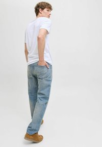 Joven con camiseta blanca y jeans azules caminando con una mano en el bolsillo trasero, mirando por encima del hombro contra un fondo blanco liso.