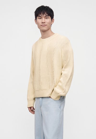 Jeune homme debout, une main dans la poche, portant un pull torsadé couleur crème et un jean bleu clair sur un fond uni.
