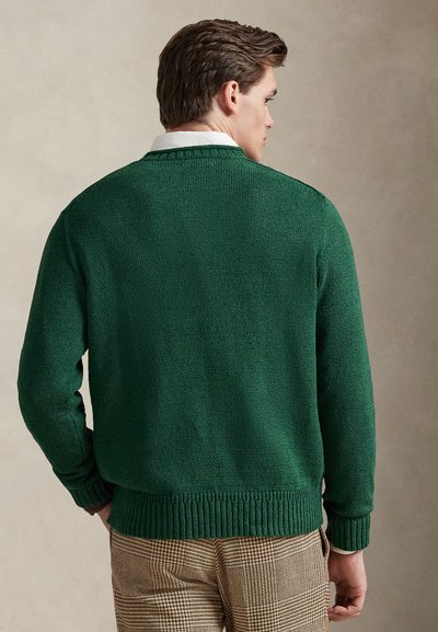 Πράσινο πουλόβερ πλεκτό με υφή, με ριμπ μανσέτες και τελείωμα, φορεμένο πάνω από μπεζ και καφέ καρό παντελόνια. Η οπίσθια όψη αναδεικνύει τον απλό σχεδιασμό.