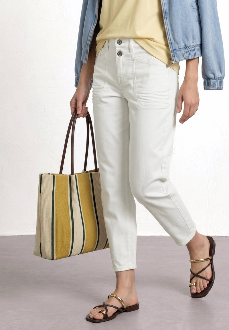Personne portant un pantalon blanc, un haut jaune, une veste bleu clair, des sandales marron avec des détails dorés, tenant un grand sac fourre-tout rayé jaune et beige.