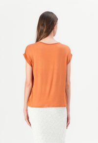 Femme aux longs cheveux bruns portant un t-shirt orange à manches courtes et une jupe blanche texturée, vue de dos.