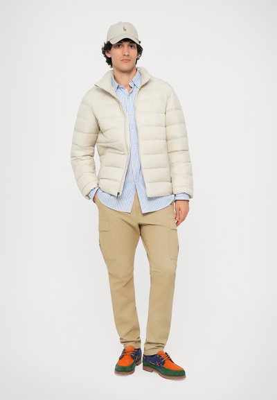 Giubbotto puffer beige chiaro, camicia a righe blu, pantaloni cargo beige e scarpe multicolori. Il giubbotto presenta un colletto alto e chiusura con zip.