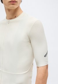 Κοντινό πλάνο ενός ατόμου που φοράει ποδηλατική μπλούζα σε ανοιχτό μπεζ με υφή, με φερμουάρ μπροστά και το κείμενο «sport» στο μανίκι.