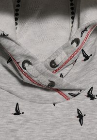 Grå tygertröja med svarta fågelmönster. Innehåller metallöglor och vita dragsn�ören, med röda stygn som accent.