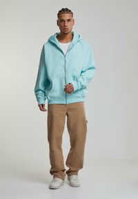 Lyseblå zip-up hoodie med kengurulommer, parret med beige bukser og hvide sneakers. Hoodien har en afslappet pasform og en blød tekstur.