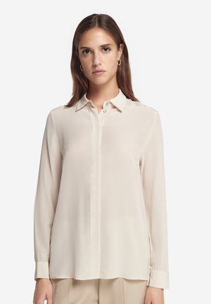 Kvinde med glat brunt hår iført en cremefarvet bluse med lange ærmer og beige bukser, stående mod en hvid baggrund.